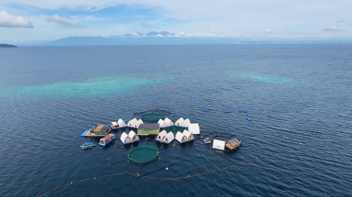 Aerial photograph of the Reef Resort in operation: white peaked glamping tents and solar panels on a floating platform between two circular fish pens, moored over a turquoise reef with service boats alongside and a mountain range on the far horizon