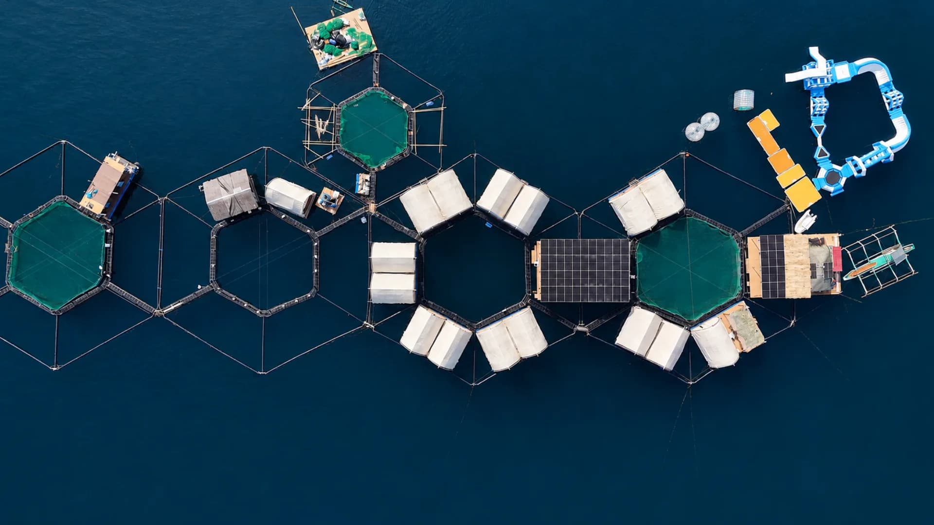 Top-down aerial photograph of the Reef Resort: hexagonal fish pens with green netting, rows of white glamping tents on floating platforms, black solar arrays, and an inflatable water park moored alongside on deep blue water
