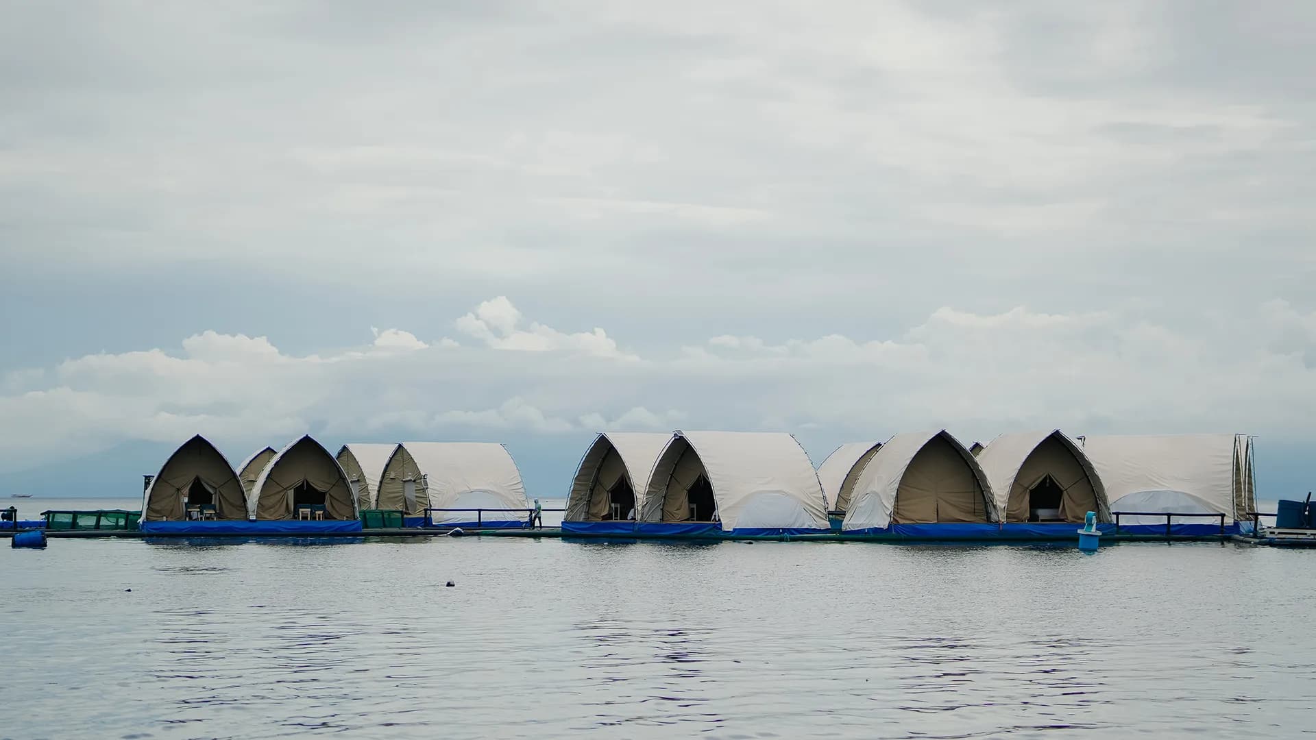 A row of peaked white Glamphouse tents on blue floating pontoons, photographed from the water under an overcast sky