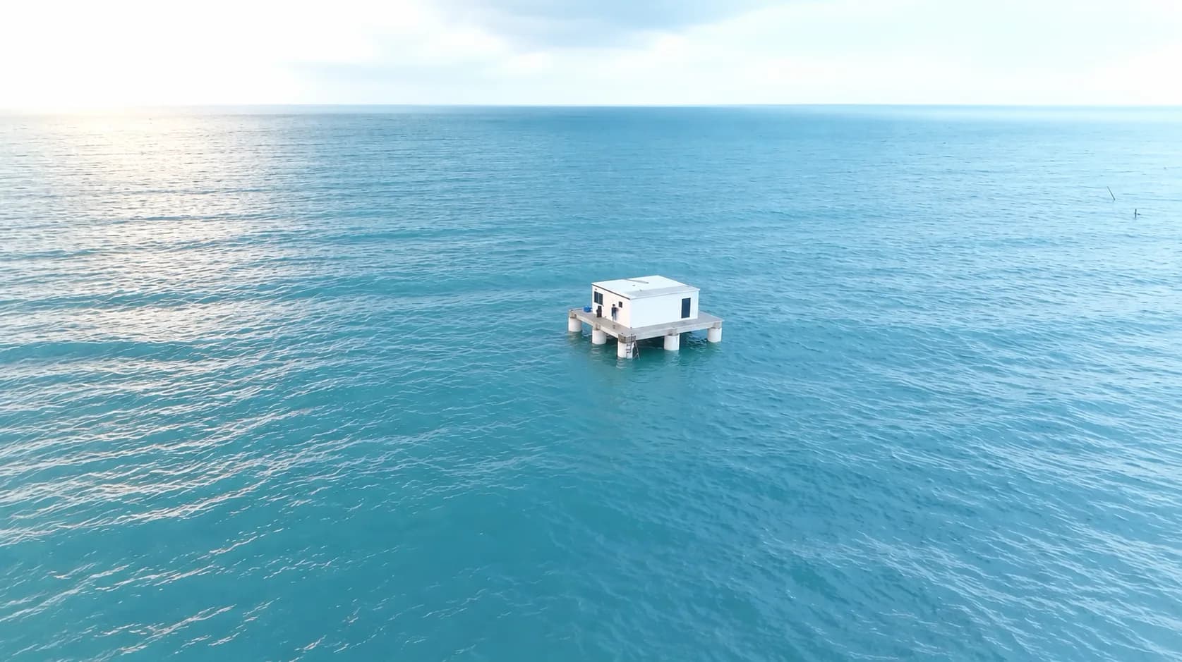 Photograph of a single ArkPad platform alone on open turquoise sea under a bright sky: a white cabin on a deck carried by cylindrical spars, with sunlight glinting off the water behind it